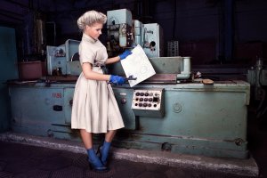 A woman in a vintage-style dress and blue gloves stands in front of an industrial machine, holding a large blueprint as if preparing for a job interview. She looks at the blueprint thoughtfully amidst the workshop's various equipment and control panels.