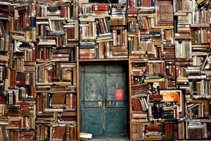 A wooden door, reminiscent of entering a job interview, is surrounded by a chaotic arrangement of old books stacked vertically and horizontally, filling the entire wall around it. A small red sign is posted on the door, and one book is positioned open to reveal its colorful pages.