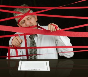 A man in a suit is tangled in excessive red tape, attempting to cut it with scissors. He looks frustrated and is struggling to reach a contract on the table in front of him, symbolizing bureaucratic obstacles.