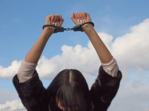 A person is shown with their hands raised above their head, wearing handcuffs. They have long dark hair and are dressed in a fur-like coat. The background, amidst a clear blue sky with a few scattered clouds, hints at the red tape that contrasts starkly with their predicament.