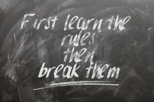 A blackboard with chalk writing that reads, "First learn the rules; then break them." The text, written in white chalk with some areas smudged, gives a rough and artistic appearance reminiscent of navigating red tape.