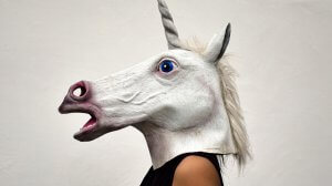 A person is wearing a realistic, full-head unicorn mask that features a white mane, a long white horn, a pink and white muzzle, and a blue eye. Dressed in black and seen from the side against a plain white background, they stand out like resume myths in the world of job hunting.