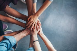 A group of diverse hands, reminiscent of the collaborative spirit of a social worker, are placed on top of each other, symbolizing unity and teamwork. The background is a smooth, gray surface. The hands include different skin tones and are seen from a top-down perspective.