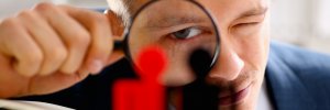 A social worker closely examines two miniature figures, one red and one black, through a magnifying glass. The scene suggests a detailed scrutiny or analysis, with the person's eye prominently focused for a clearer view.