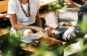 Two people having a business meeting outdoors at a wooden table. One person, a social worker, gestures with their hands near an open laptop while the other listens intently. The table holds a tablet, smartphone, and notebooks. Lush greenery surrounds the seating area.