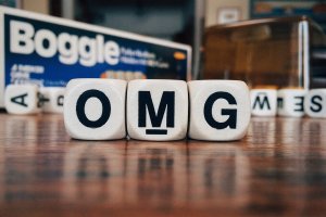 Close-up of Boggle game dice on a table spelling out "OMG" in large black letters. The Boggle game box is visible in the background along with other scattered dice, resembling a scene from a social worker's office during an engaging session. The surface of the table shows a wood grain pattern.