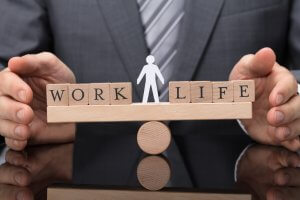 A person in a suit places their hands around a balance scale made of wooden blocks with the words "WORK" and "LIFE" on either side. A small human figure stands between the words, symbolizing the delicate act of job surviving within work-life balance.