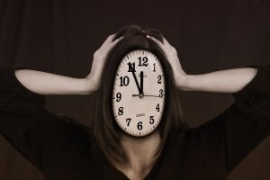 A person with long hair holding their head in their hands has a clock for a face, displaying the time as 11:55. This surreal image symbolizes job-related stress or anxiety about surviving in a demanding environment. The dark background intensifies the overall atmosphere.