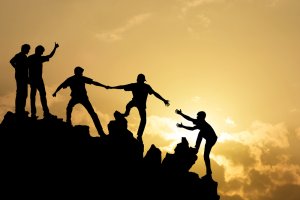 Silhouetted against a golden sunset, a team of four people on a rocky peak help a fifth person climb to the top. The scene captures themes of teamwork, support, and achievement, with the sun casting a warm glow through scattered clouds in the background.