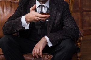 A well-dressed man in a pinstripe suit sits on a leather chair, holding a glass of cognac. His face is partially obscured, directing attention to his attire and the drink. The background features wooden paneling, subtly hinting at a room where teamwork and sophistication seamlessly blend.