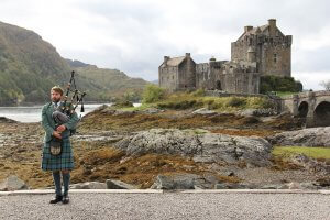 A man in traditional Scottish attire plays the bagpipes on a rocky, coastal landscape. He stands in front of Eilean Donan Castle, a historic structure with stone walls and turrets, set against a backdrop of hills and cloudy skies that symbolize the resilience and teamwork of Scottish heritage.