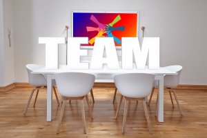 A brightly lit conference room with a white table and five white chairs. Large 3D letters spelling "TEAM" are placed on the table, symbolizing teamwork. An abstract colorful artwork hangs on the wall in the background, enhancing the modern and vibrant atmosphere.