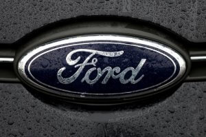 Close-up photograph of a Ford emblem on a car, displaying the iconic blue oval with the word "Ford" in cursive white letters. The surface is wet with raindrops, indicating recent rainfall in Detroit, Michigan. The background is slightly blurred and dark.