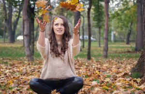 A young woman with long curly hair sits cross-legged on the ground in a park filled with fallen leaves. She is wearing a beige sweater and black pants, joyfully tossing maple leaves into the air. Trees and greenery frame this serene moment in the background.