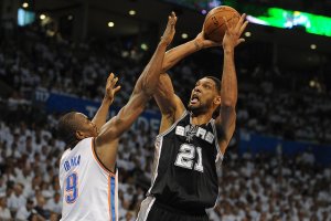 Two basketball players in action during a game. One player wearing a black jersey numbered 21 is attempting a jump shot, while a player in a white jersey numbered 9 tries to block the shot. The background captures the authenticity of the moment with a crowd of spectators and a partially visible scoreboard.
