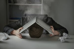 A person in a suit leans their head against a desk with an open laptop resting on their head like a roof. Smoke, likely from burnout, billows from the laptop. A computer mouse and a crumpled piece of paper lie on the desk, while shelves are visible in the background.