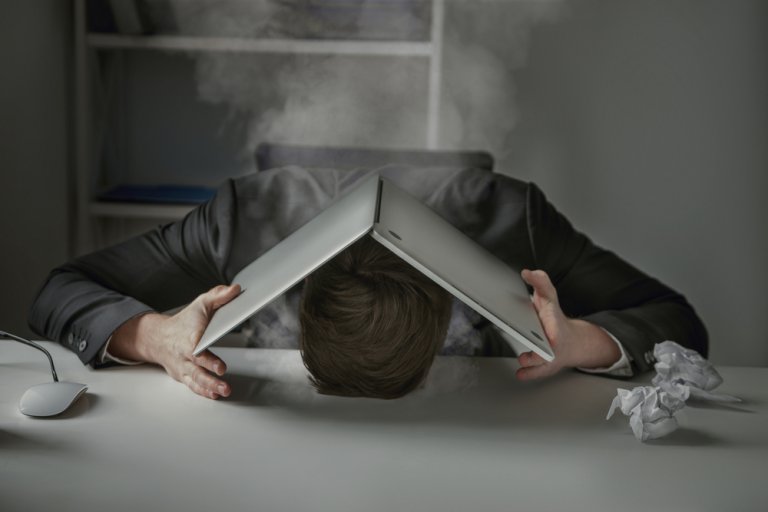 A person in a suit leans their head against a desk with an open laptop resting on their head like a roof. Smoke, likely from burnout, billows from the laptop. A computer mouse and a crumpled piece of paper lie on the desk, while shelves are visible in the background.