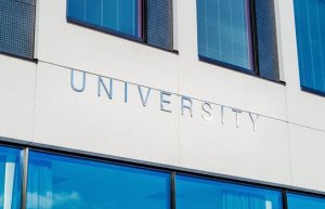Exterior view of a building with large windows on the upper floors and the word "UNIVERSITY" displayed prominently on the facade. The windows reflect a clear blue sky, indicating a bright day. The overall design is modern and minimalistic, reflecting the cutting-edge focus on cybersecurity education inside.