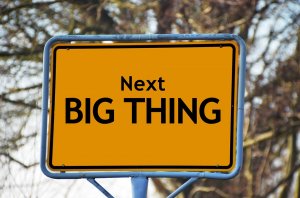 A yellow road sign with bold black text that reads "Next BIG THING" stands against a background of blurred, leafless trees. The sign is set in a metal frame and suggests an upcoming exciting or significant development in the field of cybersecurity.