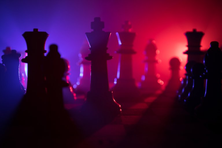 Silhouetted chess pieces standing on a chessboard are illuminated by dramatic lighting, with a blue light on the left and a red light on the right, creating an intense, high-contrast ambiance reminiscent of a strategic development plan.