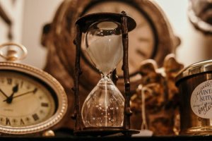 A close-up of an hourglass surrounded by vintage items. To the left is an antique clock with a white face and gold frame. In the background, a blurred circular clock adds depth. To the right, a label on a container reads "Vintage Suit" amidst other indistinct items, like pages of an old development plan.