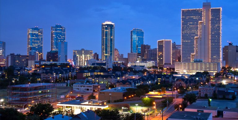 A nighttime cityscape of Fort Worth, Texas, featuring tall, illuminated skyscrapers with lights outlining their edges against a deep blue sky. Smaller buildings and houses populate the foreground, with streetlights casting a warm glow on the roads below.