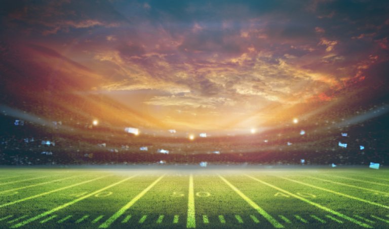A football field is illuminated under a dramatic sunset, with vibrant red and orange hues in the sky. The stadium seating in the background is filled with spectators cheering on their teams, creating a lively atmosphere. The field markings and yard lines are clearly visible.