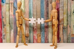 Two wooden mannequin figures stand facing each other, each holding a puzzle piece that fits together. The background consists of wooden panels painted in various colors, creating a vibrant backdrop that highlights the essence of teamwork.