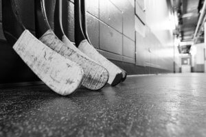 Black and white image of three hockey sticks leaning against a wall in a narrow, dimly lit hallway. The sticks have worn, taped blades and rest on a textured floor. The scene evokes a sense of anticipation before the game, brimming with the promise of success.