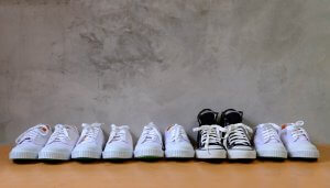 A row of eight pairs of sneakers is seen on a wooden surface against a textured gray wall. Seven pairs of the sneakers are white, and one pair is black with white laces and details, standing out like experienced job seekers among newcomers.