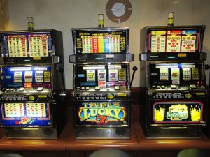 Three colorful slot machines stand side by side in a casino, doing their job to entice players with vibrant graphics. One features the "Triple Lucky 7" game, while another showcases a "Super Reel" game. The dimly lit area around the machines adds to the allure and excitement.