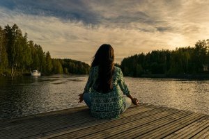 A person sits cross-legged on a wooden dock facing a serene lake surrounded by trees. The sky is partly cloudy with warm colors of the sunset reflecting on the water. The individual appears to be meditating, perhaps finding solace after a long day at their job, adding to the peaceful atmosphere of the scene.