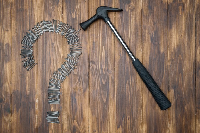 A hammer lies on a wooden surface next to a large question mark made out of nails, evoking a sense of mystery akin to an interview. The wood has a natural, dark finish with visible grain patterns. The hammer has a black handle and a silver neck.