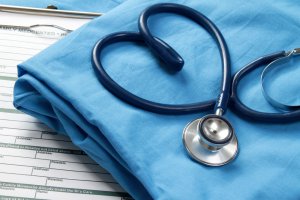 A blue stethoscope rests atop a folded blue medical gown, forming a heart shape with its tubing. Underneath, a medical form on a clipboard is partially visible. The scene evokes a nursing or healthcare setting.