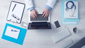 A nursing professional in a white coat types on a laptop at a desk. The desk has medical equipment like a stethoscope, thermometer, notepads, glasses, keyboard, smartphone, and a cup of pens. The setup suggests attentive care and dedication in the medical field.
