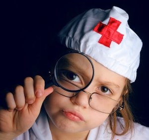 A young child dressed as a nurse is holding a magnifying glass up to her eye, which is enlarged behind the lens. She is wearing a white nurse's cap with a red cross on it and is looking intently at the camera with a playful expression, embodying her nursing aspirations even at such a tender age.