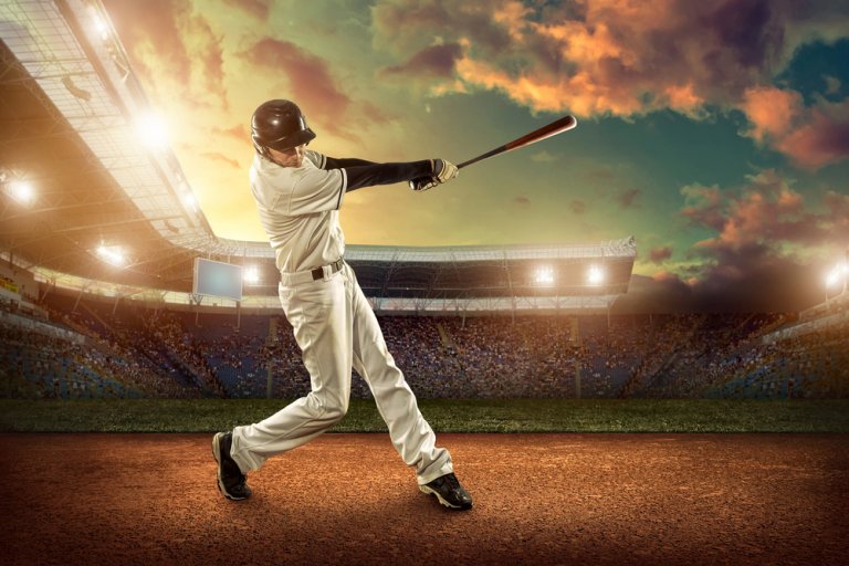 A baseball player in a white uniform swings a bat during a game in a stadium at sunset. The sky is a mix of orange, yellow, and blue hues, and the stadium lights are on. The player is captured mid-swing, focusing intensely on hitting the ball as questions linger about the game's outcome.