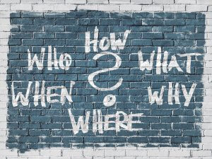A brick wall painted with a teal background features the words "WHO," "HOW," "WHAT," "WHEN," "WHY," and "WHERE" in large, white, bold letters arranged around a central, large question mark—inviting endless questions.