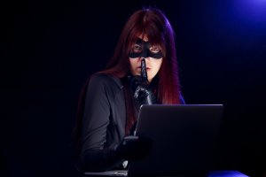 A person with long red hair and a black mask covers their mouth with a gloved finger in a shushing gesture while looking at a laptop, possibly on a job search. The background is dark with a purple light shining from the top right corner.
