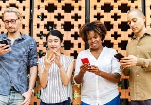Four people are standing against a decorative brick wall, each looking at their smartphone. Dressed casually and focused on their screens, they seem engrossed in a job search. The wall features a geometric pattern.