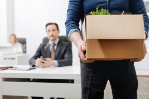 A person in business attire holds a cardboard box with personal items, including a potted plant, standing in the foreground. In the background, two people are seated at desks, observing the scene of someone leaving their job. The room appears to be an office.