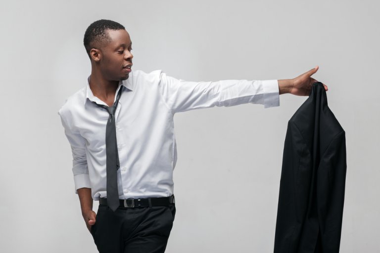 A man wearing a white dress shirt and black tie holds a black jacket in his outstretched right hand, as if contemplating leaving your job. His left hand is in his pocket, and he is looking towards the jacket. The background is plain and white.
