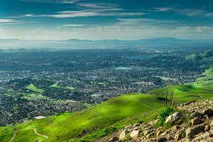 A panoramic view from a hilltop, overlooking the sprawling cityscape of Silicon Valley surrounded by lush green hills. The sky is clear with a few scattered clouds, and a large body of water is visible in the distance. The landscape is bathed in bright sunlight.