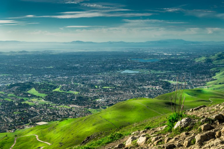 A panoramic view from a hilltop, overlooking the sprawling cityscape of Silicon Valley surrounded by lush green hills. The sky is clear with a few scattered clouds, and a large body of water is visible in the distance. The landscape is bathed in bright sunlight.