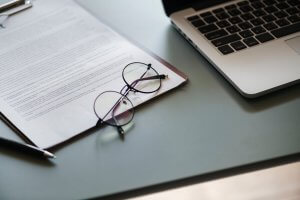A pair of glasses rests on top of a clipboard with papers containing text, including a cover letter. A pen lies next to the clipboard on a gray surface. To the right, a partial view of an open laptop is visible.