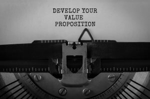 Close-up of a vintage typewriter diligently typing the words "DEVELOP YOUR VALUE PROPOSITION" on a sheet of paper. The metal type bars and paper holder are clearly visible. The text is perfectly centered on the page, and the entire image radiates with classic black-and-white charm.