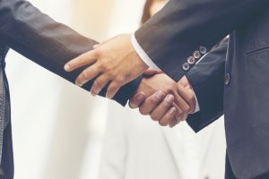 Close-up of two people in formal business attire shaking hands, symbolizing a value proposition or agreement. A person in a blurred white outfit stands in the background, slightly out of focus. The image conveys professionalism and partnership.