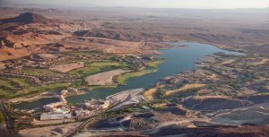 Aerial view of a desert landscape featuring a large lake surrounded by green patches of vegetation and buildings. The area includes golf courses, residential areas, and resorts. Rugged, barren hills and mountains extend into the background under a clear sky.