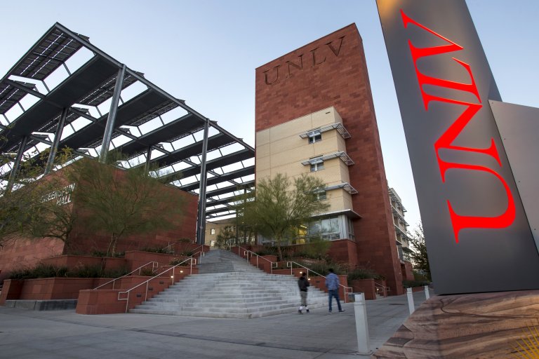 Two people walk in front of a modern building with "UNLV" prominently displayed on both a large red vertical sign and the building itself. The structure features a staircase, trees, and a partially constructed framework under a clear blue sky.