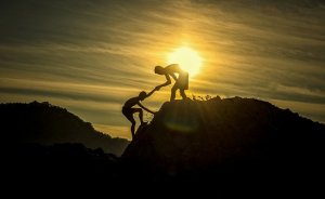 Two silhouetted figures are climbing a rocky hill at sunset, embodying the strength of relationships. One person on top extends a hand to help the other up. The background sky is filled with golden hues as the sun is setting, casting a warm glow over the scene.
